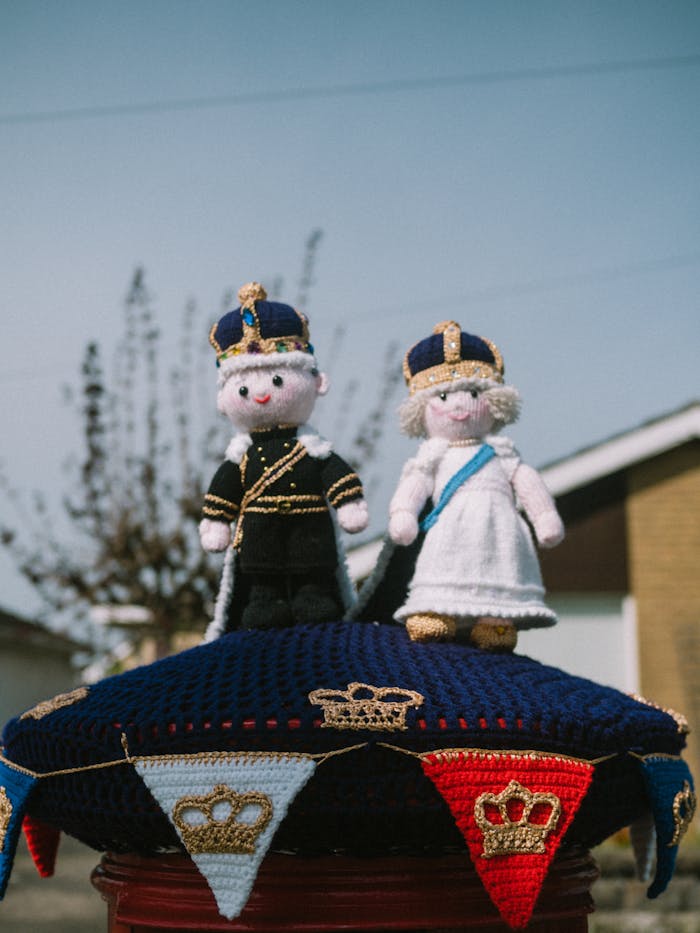 Charming royal-themed yarn dolls on a colorful knitted display outdoors under clear skies.